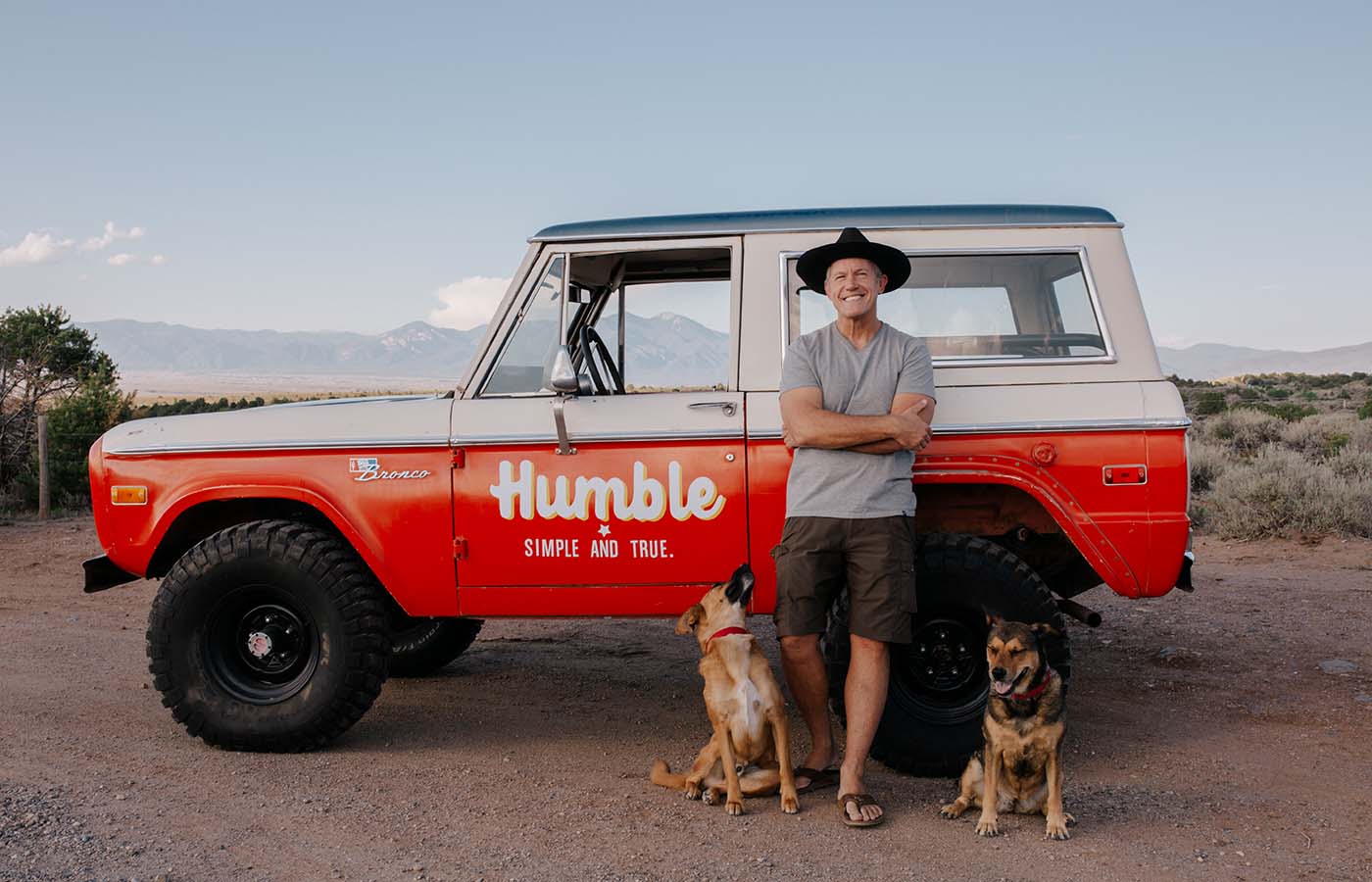 Man in hat with two dogs in front of red and white bronco truck