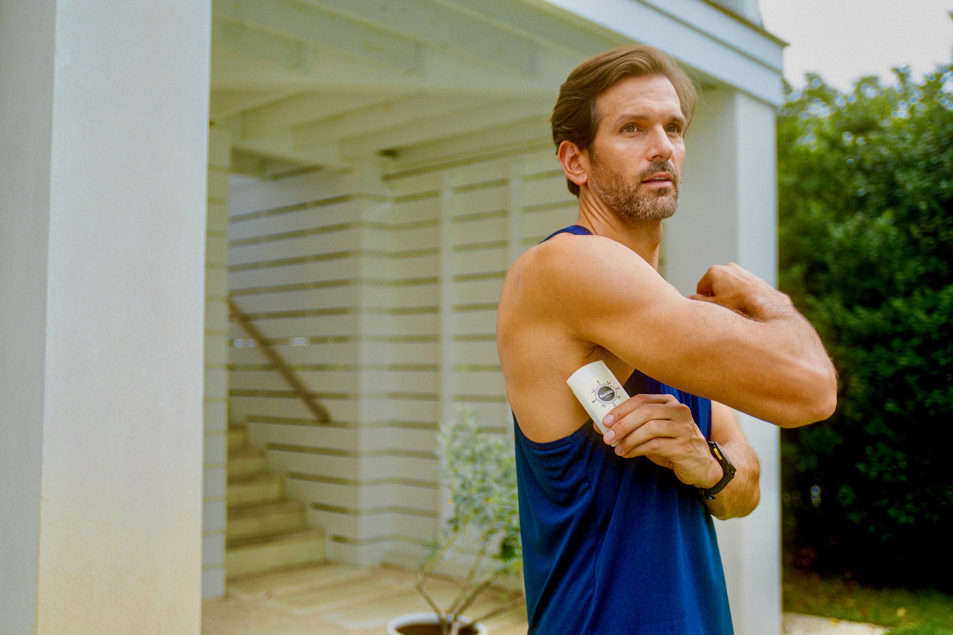 A man in a blue athletic tank top stretching his arm while holding a white bottle, standing outdoors in front of a modern house with columns and greenery.