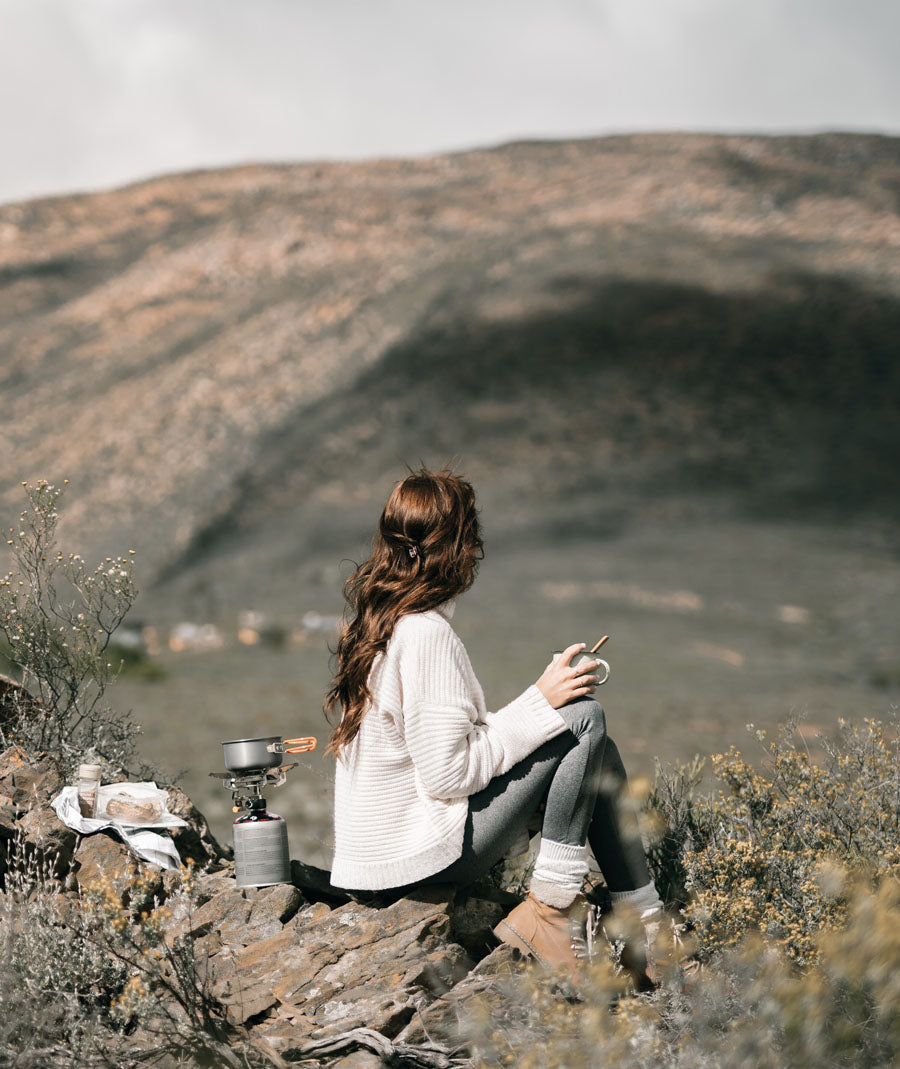 a woman sits on a rock with a camp stove looking into the distance