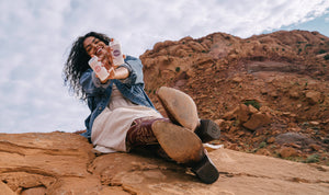 a women in a jean jacket and cowboy boots sitting in the desert holding sticks of deodorant 