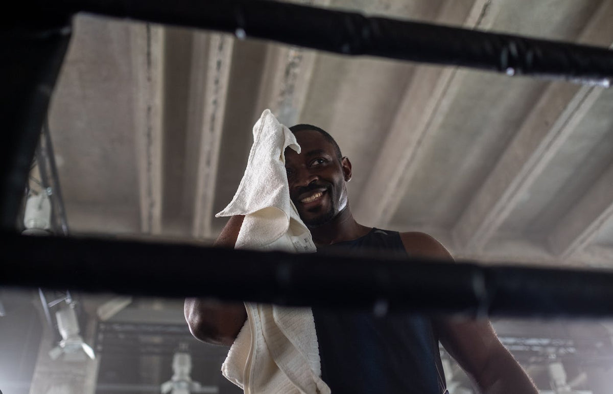 Low-angle view of a man wiping sweat with a white towel, smiling in an industrial gym seen through ropes