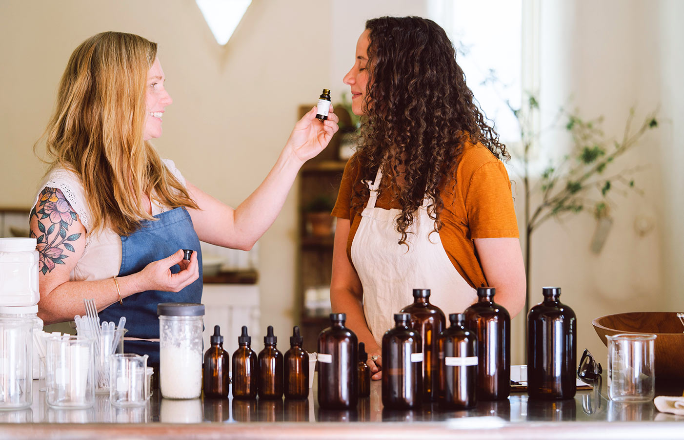 Two women in aprons sampling a botanical fragrance in a workshop with amber bottles and glassware