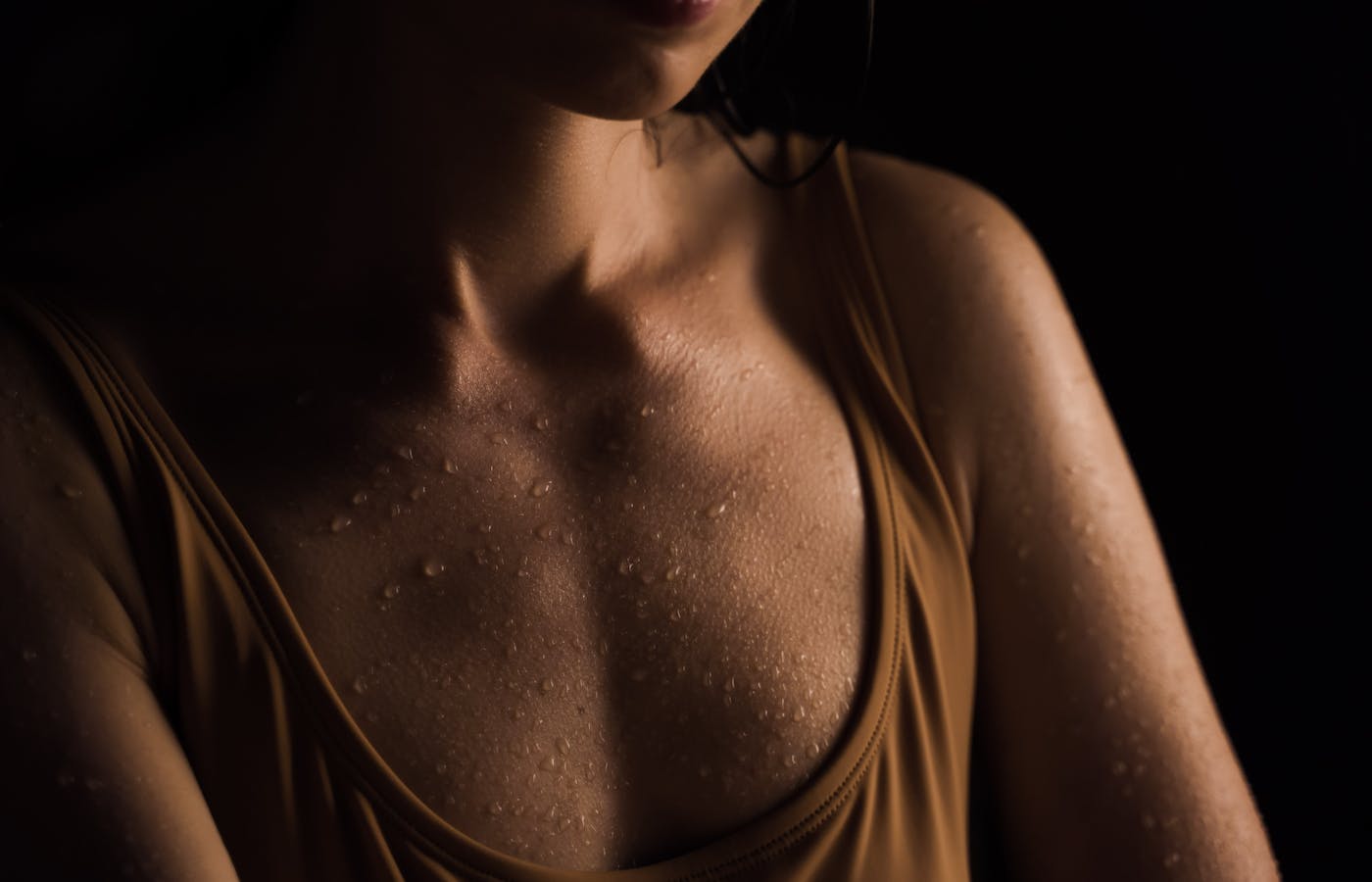 Close-up of a person’s chest and shoulders showing beads of sweat on skin while wearing a tan tank top against a dark background