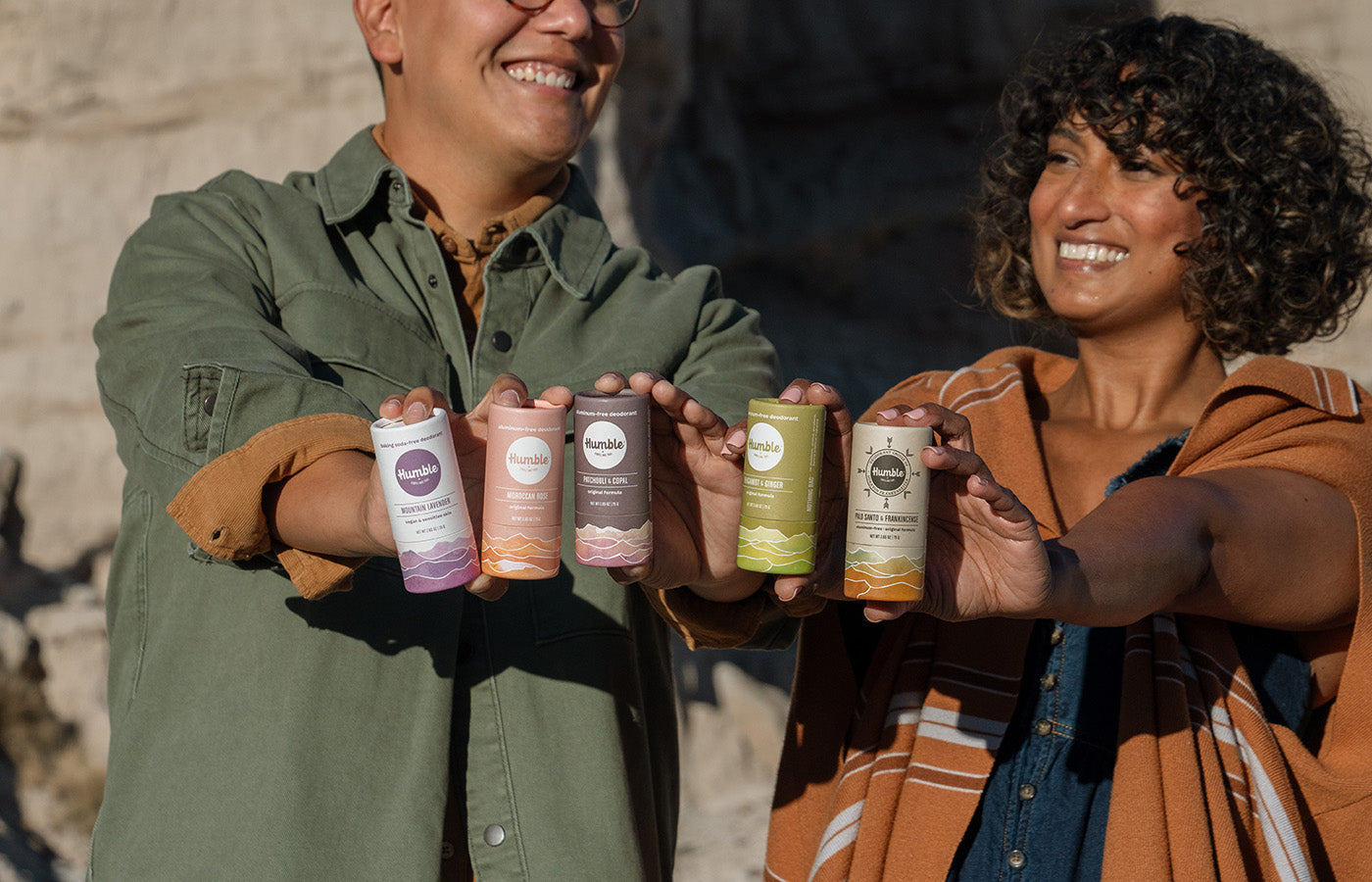 Two people holding five Humble plastic-free deodorant sticks in paper tubes outdoors against a rocky background