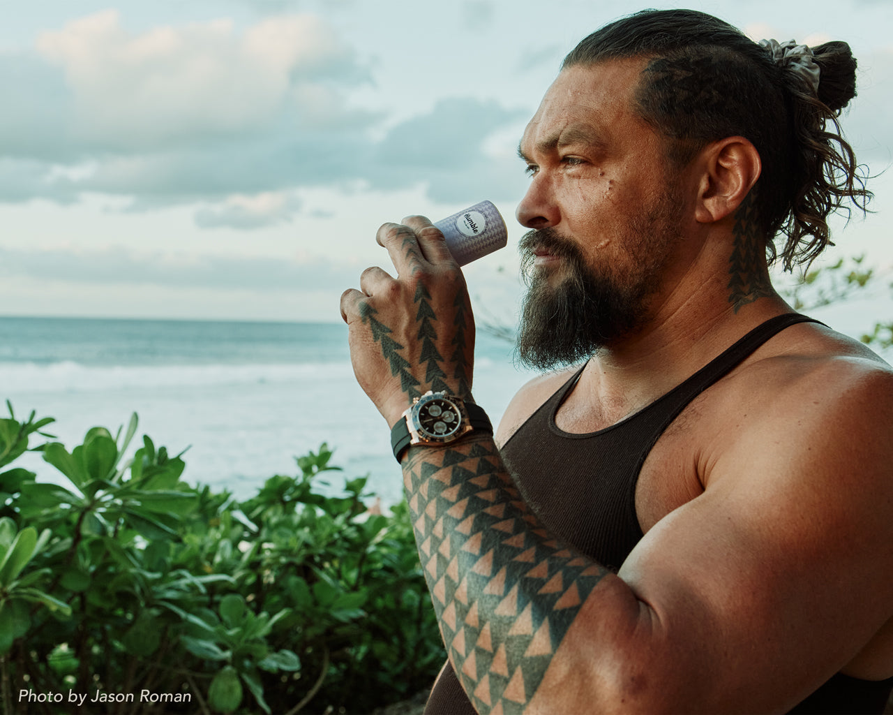Tattooed man smelling a Humble deodorant stick by the ocean with green foliage and cloudy sky in the background