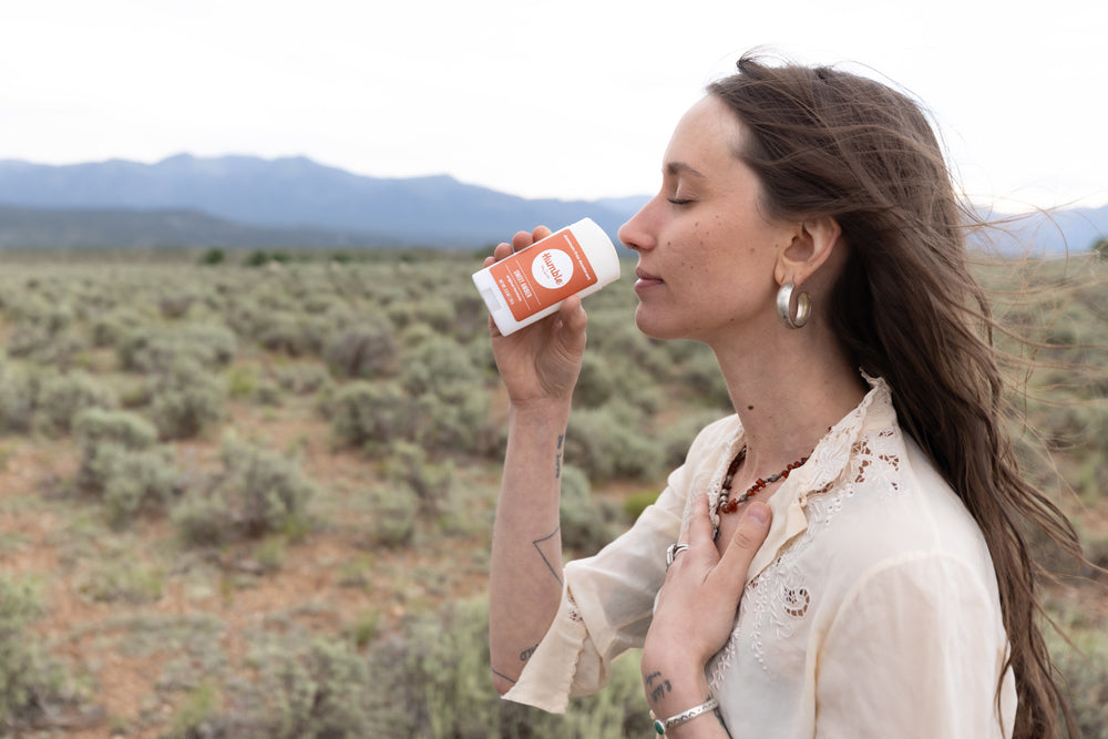 Woman smelling a Humble-branded orange-labeled scent stick in a windswept sagebrush landscape with distant mountains