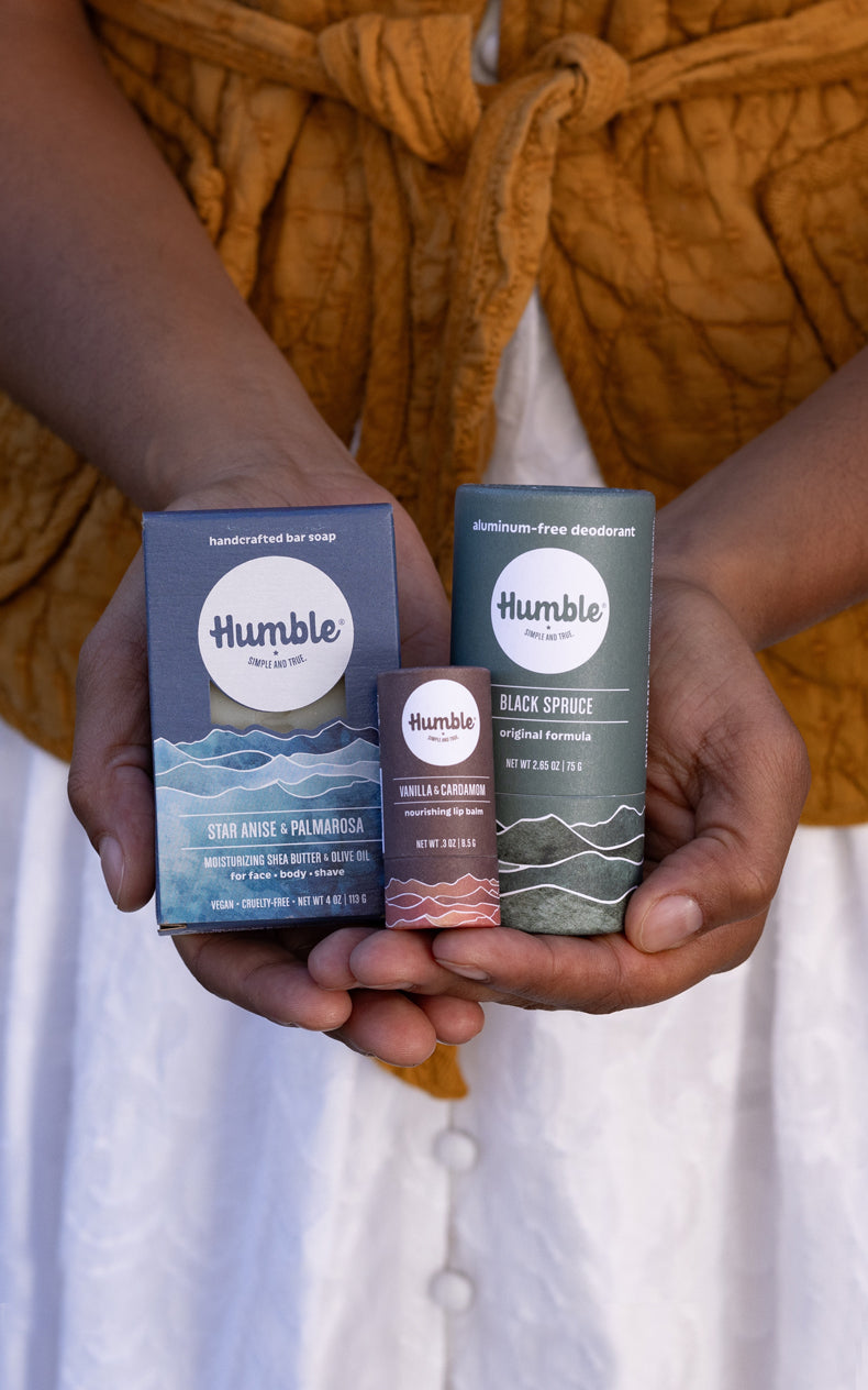 Hands holding three Humble products: Star Anise and Palmarosa soap, Vanilla and Cardamom lip balm, Black Spruce deodorant