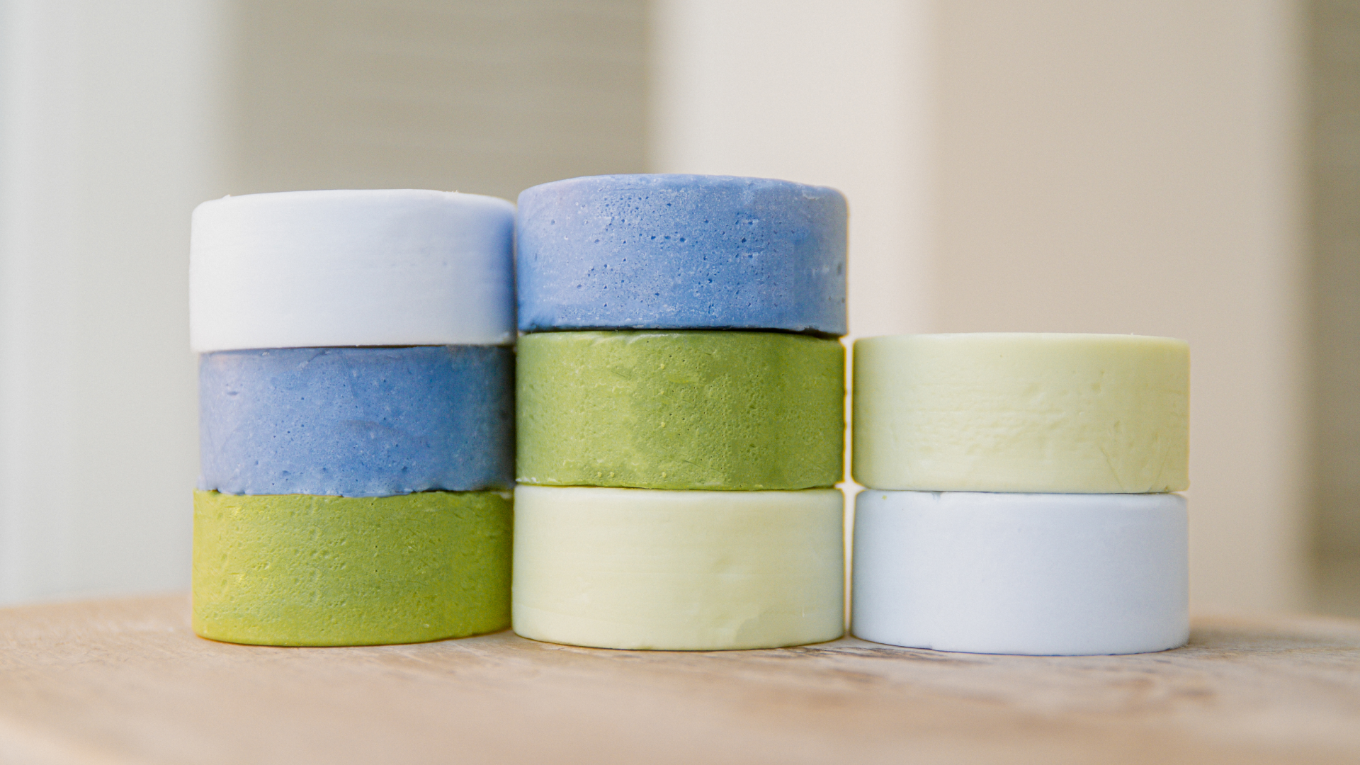 Close-up of stacked round shampoo bars in blue, green, and pale yellow showing smooth, slightly textured surfaces on a blurred neutral background