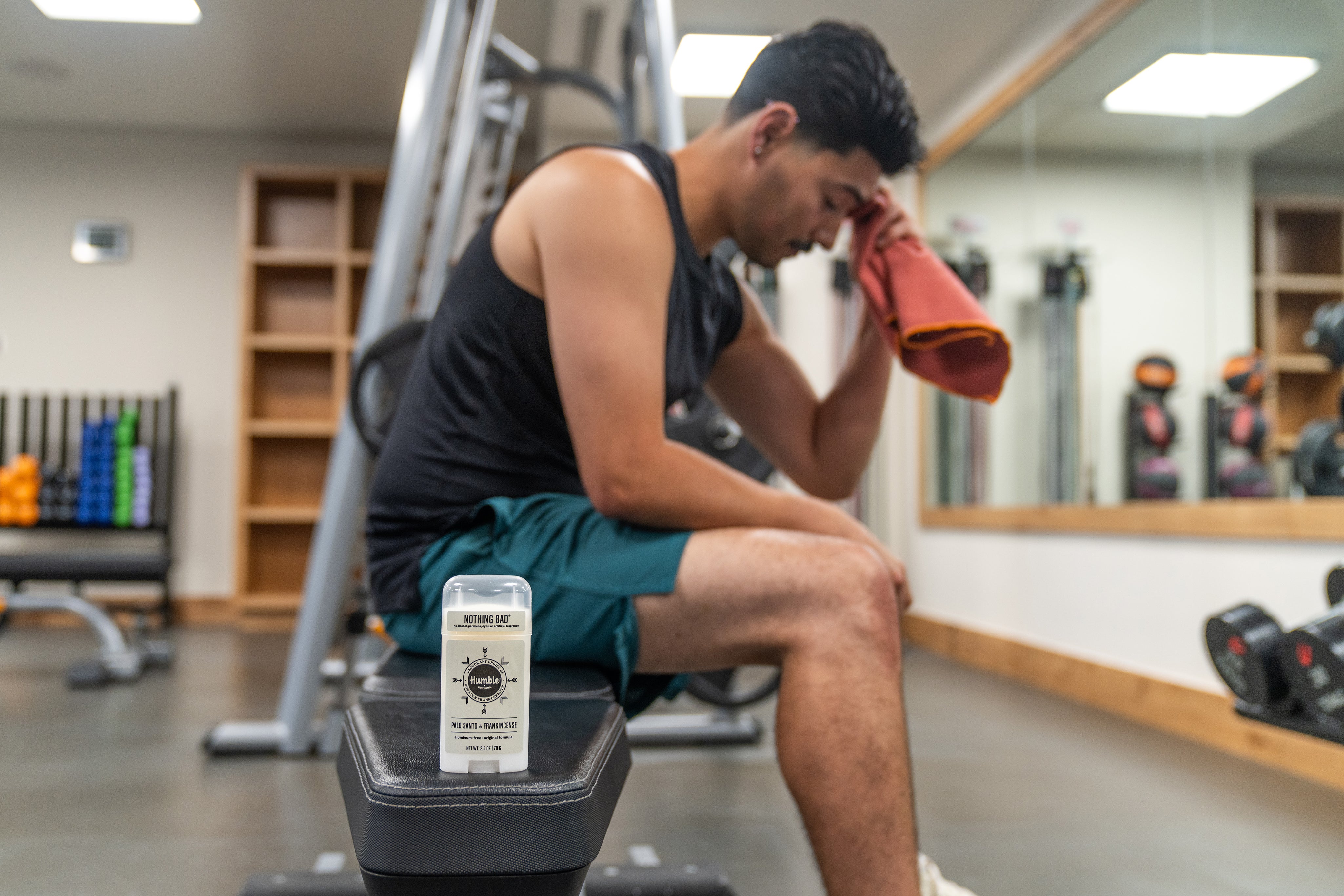 Man in gym rubbing sweat off face with deodorant on weight bench in front of him