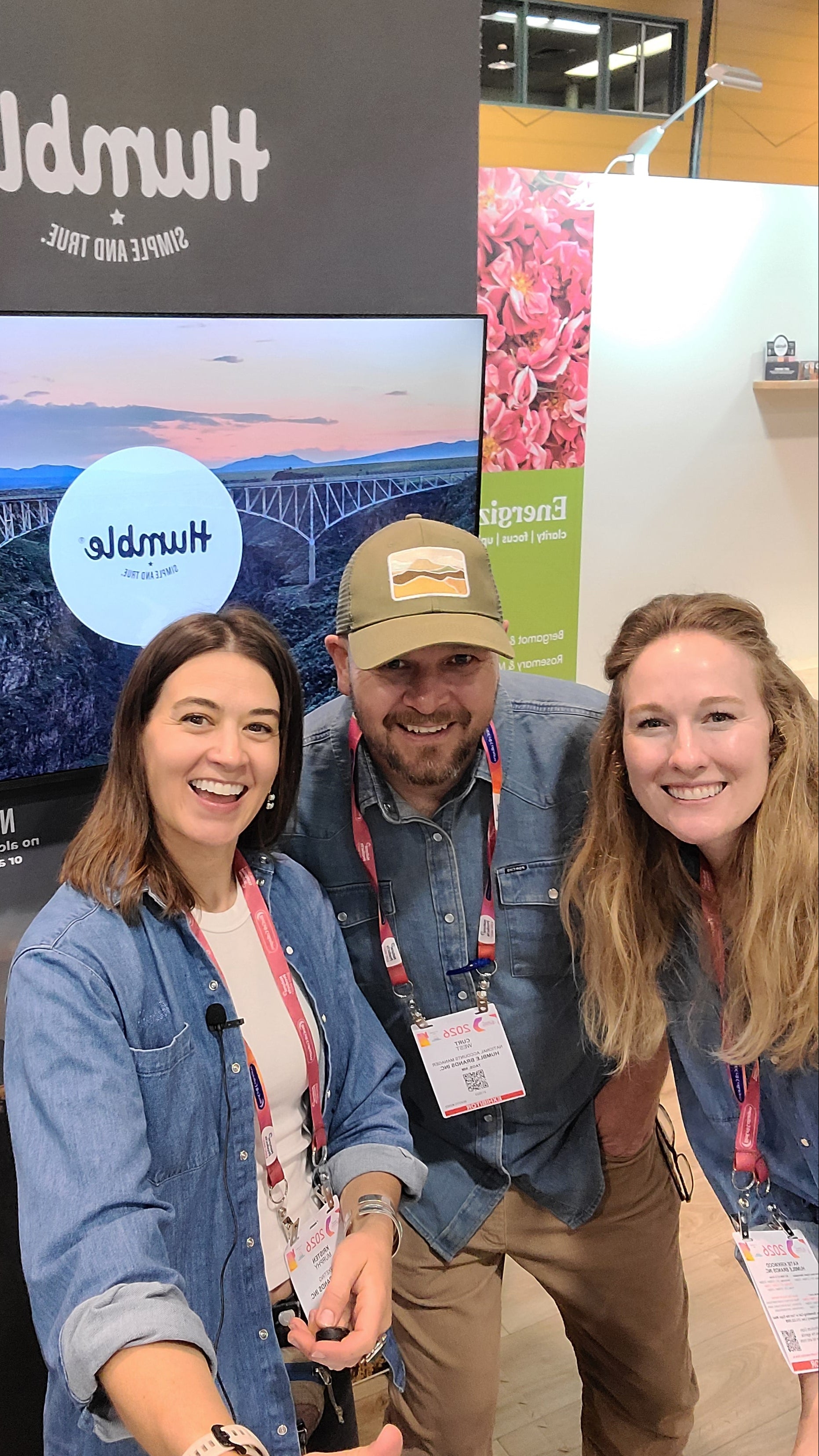 Three Humble team members smiling for a selfie at their trade-show booth with the Humble logo visible on a screen behind them
