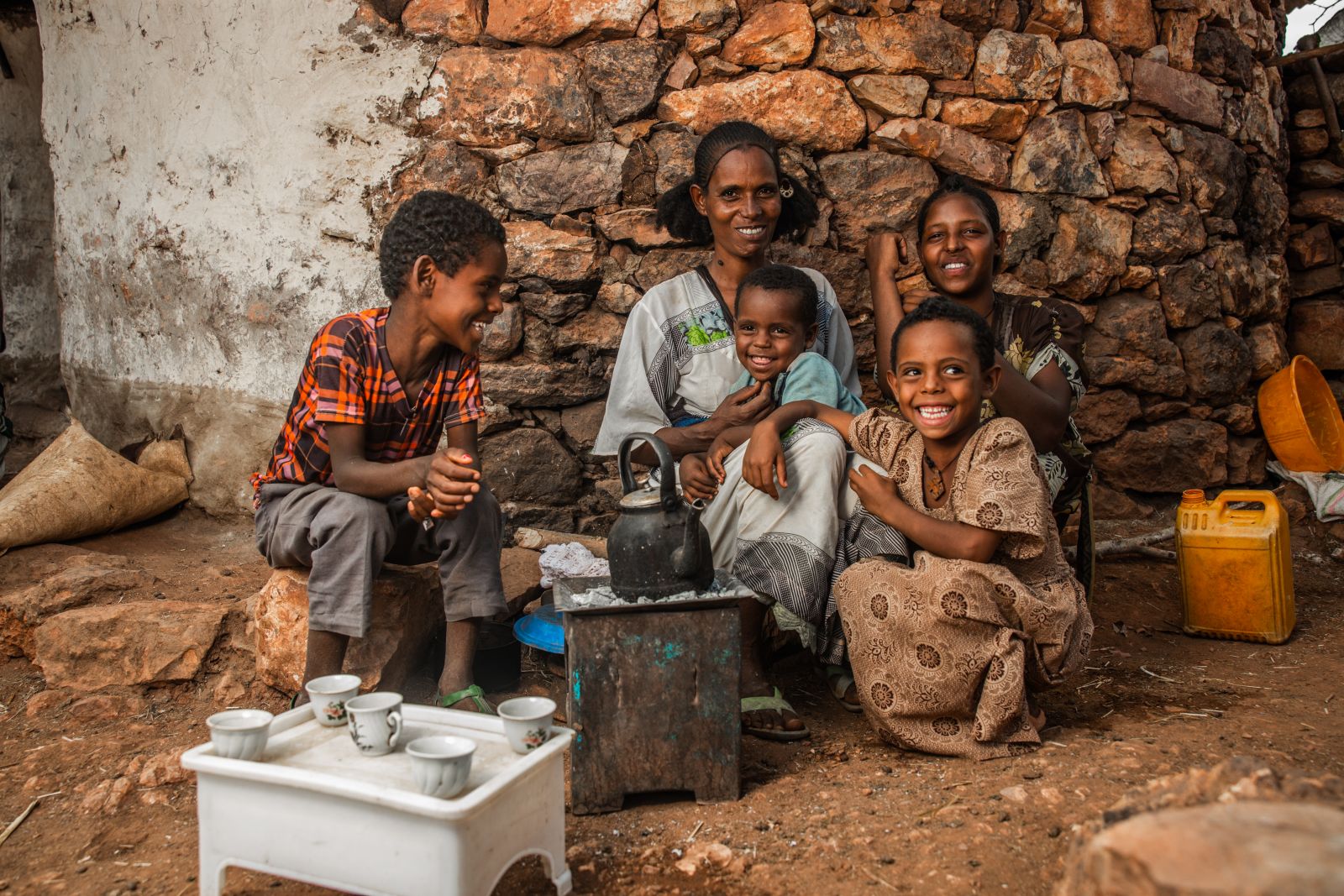a smiling family sits on the ground surrounding a tea kettle and tea cups 