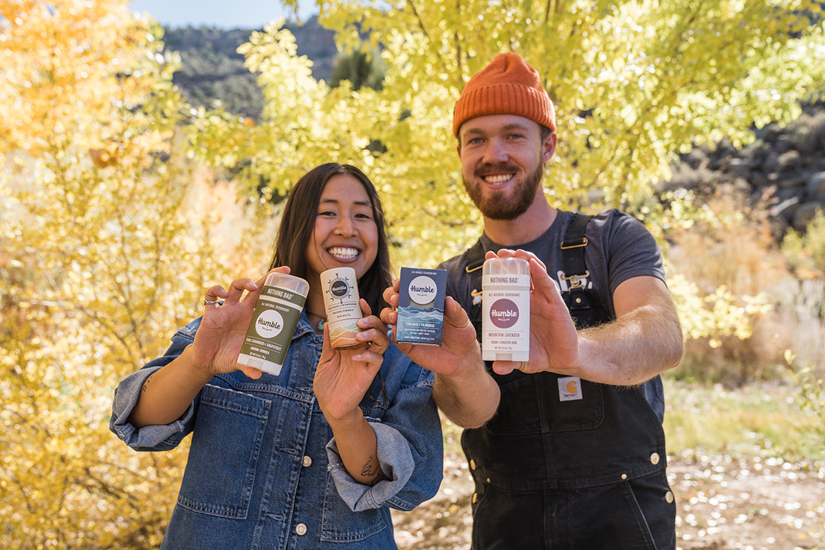 two people smiling on an autumn day and holding up soap and deodorants 
