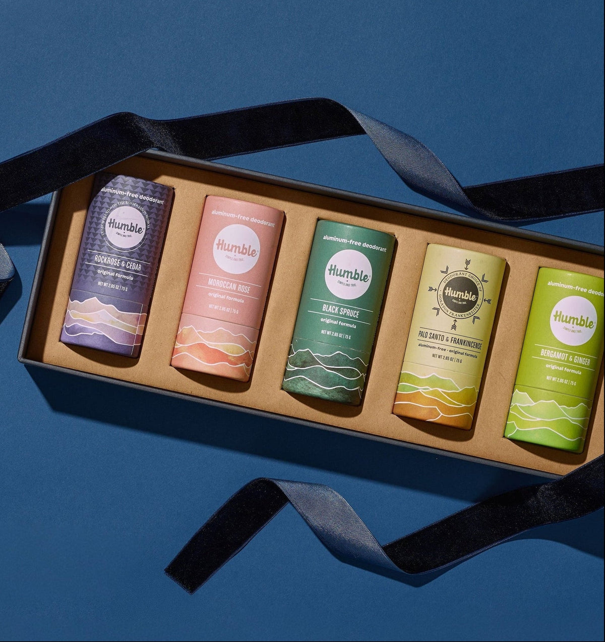 A boxed set of five Humsble personal care sticks arranged diagonally in a kraft paper tray with a black ribbon, photographed on a solid blue background.