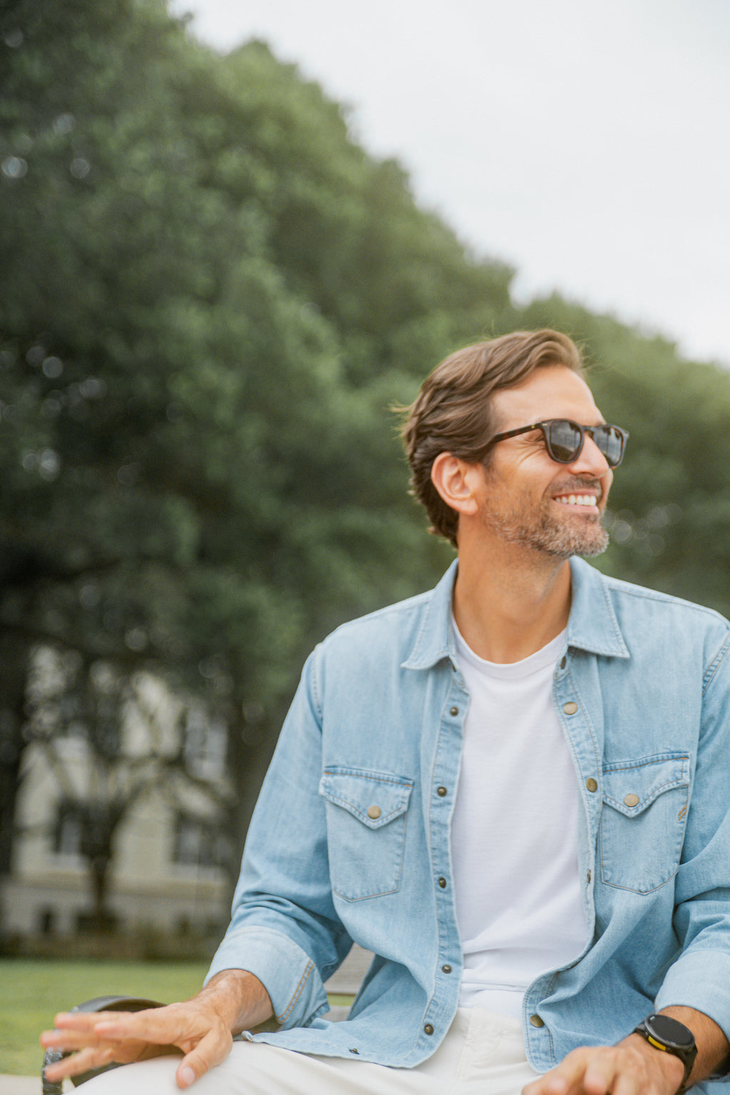 Smiling man wearing sunglasses and a light denim shirt sitting outdoors on a bench with trees in the background