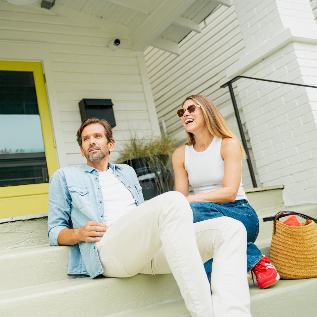 Man and woman sitting on a porch with a yellow door and white house.