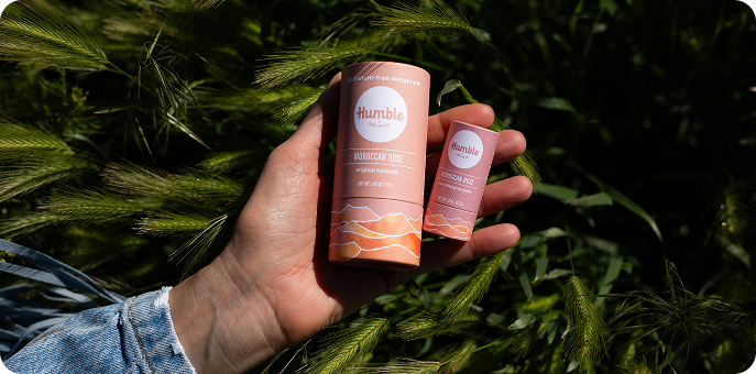 Close-up of hand holding two Humble Brands deodorant sticks labeled Moroccan Rose, showing full and travel sizes against a grassy background