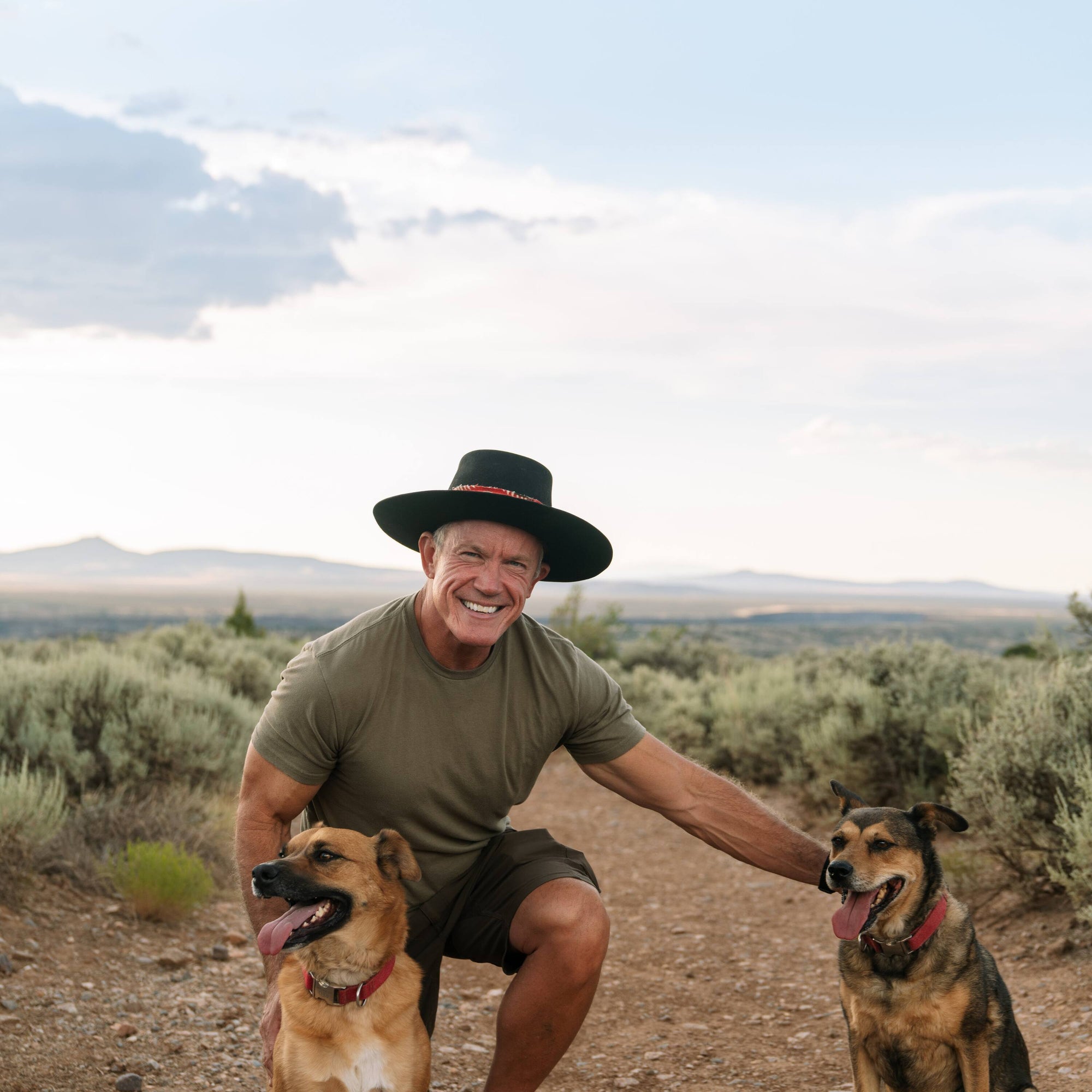 Man in a hat with two dogs on a dirt path in a desert landscape