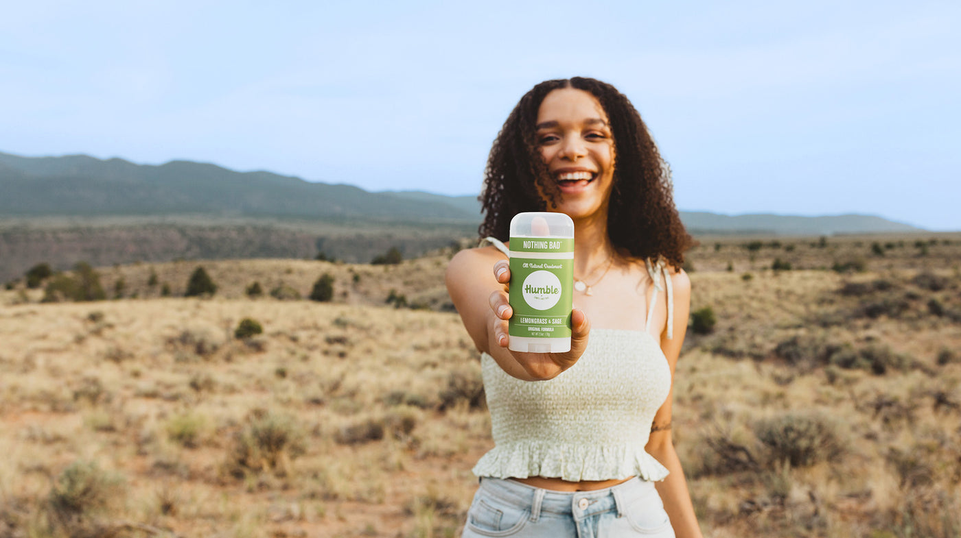 a smiling woman stands in the desert while holding a stick of deodorant out 