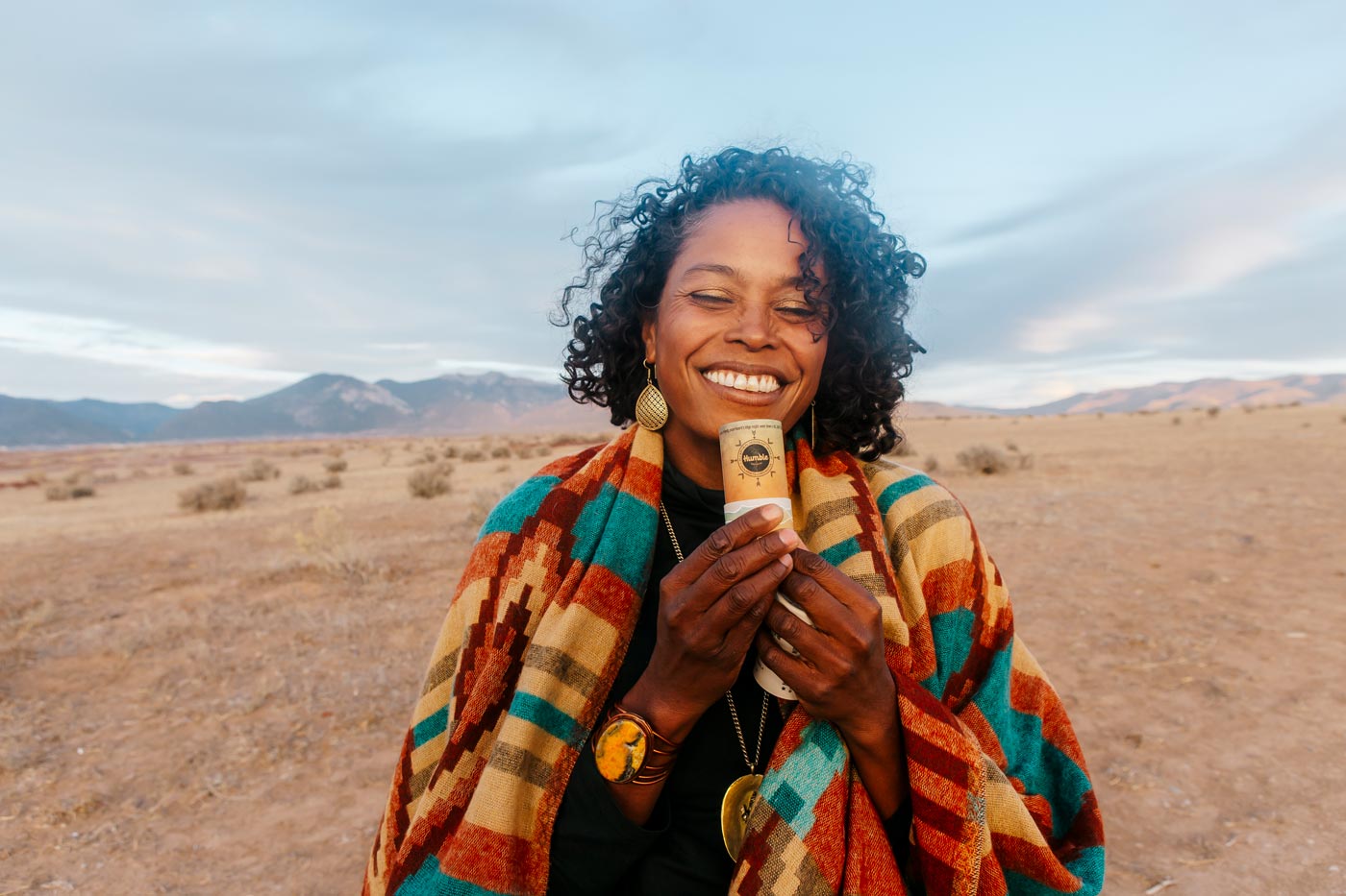 a smiling woman in a colorful jacket holds a deodorant stick while standing in the desert 