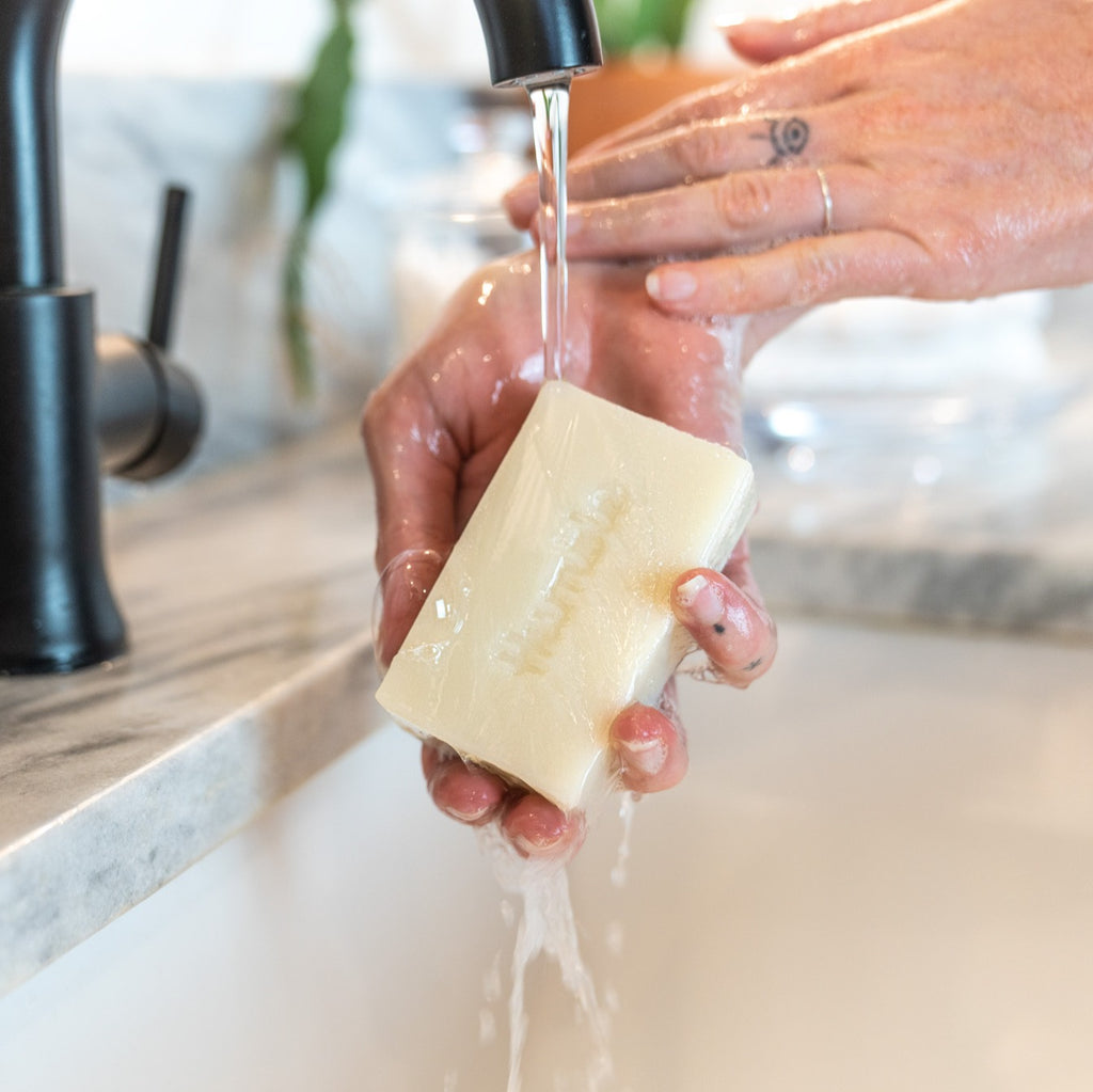 Hands rinsing a rectangular cold process bar soap embossed with Humble under a running faucet over a marble sink
