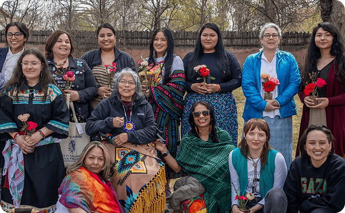 Group of women, some traditional and others in modern clothing, holding flowers outdoors.