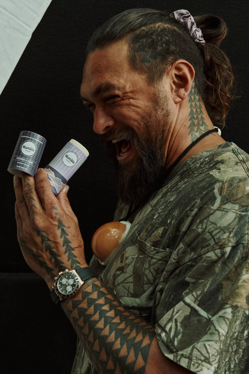 Man with tattoos laughing while holding two Humble deodorant paperboard sticks against a dark background