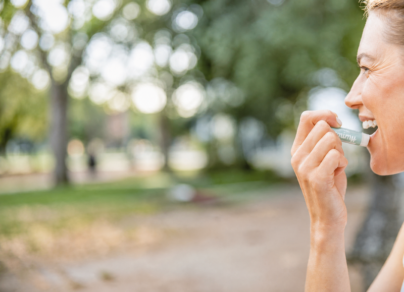 A woman in profile outdoors, holding a small white and green device to her mouth. The background is a softly blurred park with trees and a path.