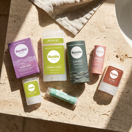 A flat-lay arrangement of six Humble brand personal care products on a beige stone surface with natural sunlight and shadows. The products include a purple box of Moroccan Spice body butter, a green and white Nothing Bag deodorant stick, a dark green Black Spice deodorant stick, a pink Moroccan Spice lip balm, a brown body butter stick, and a mint green lip balm tube.