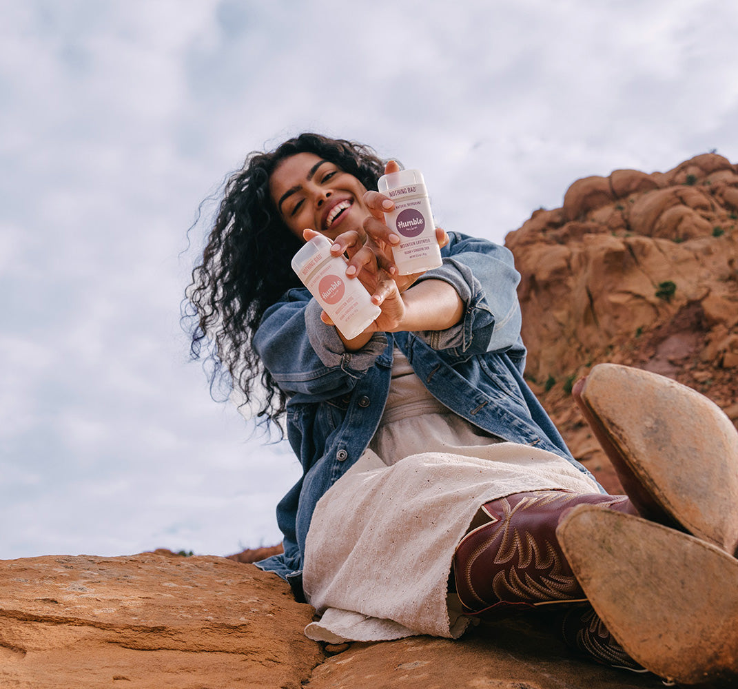 a smiling woman holds deodorant while sitting in the desert 