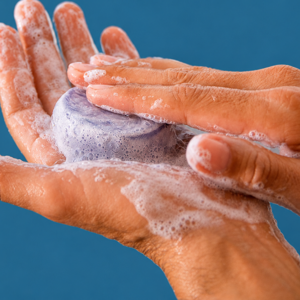 Close-up of soapy hands washing a purple bar of soap against a solid blue background.
