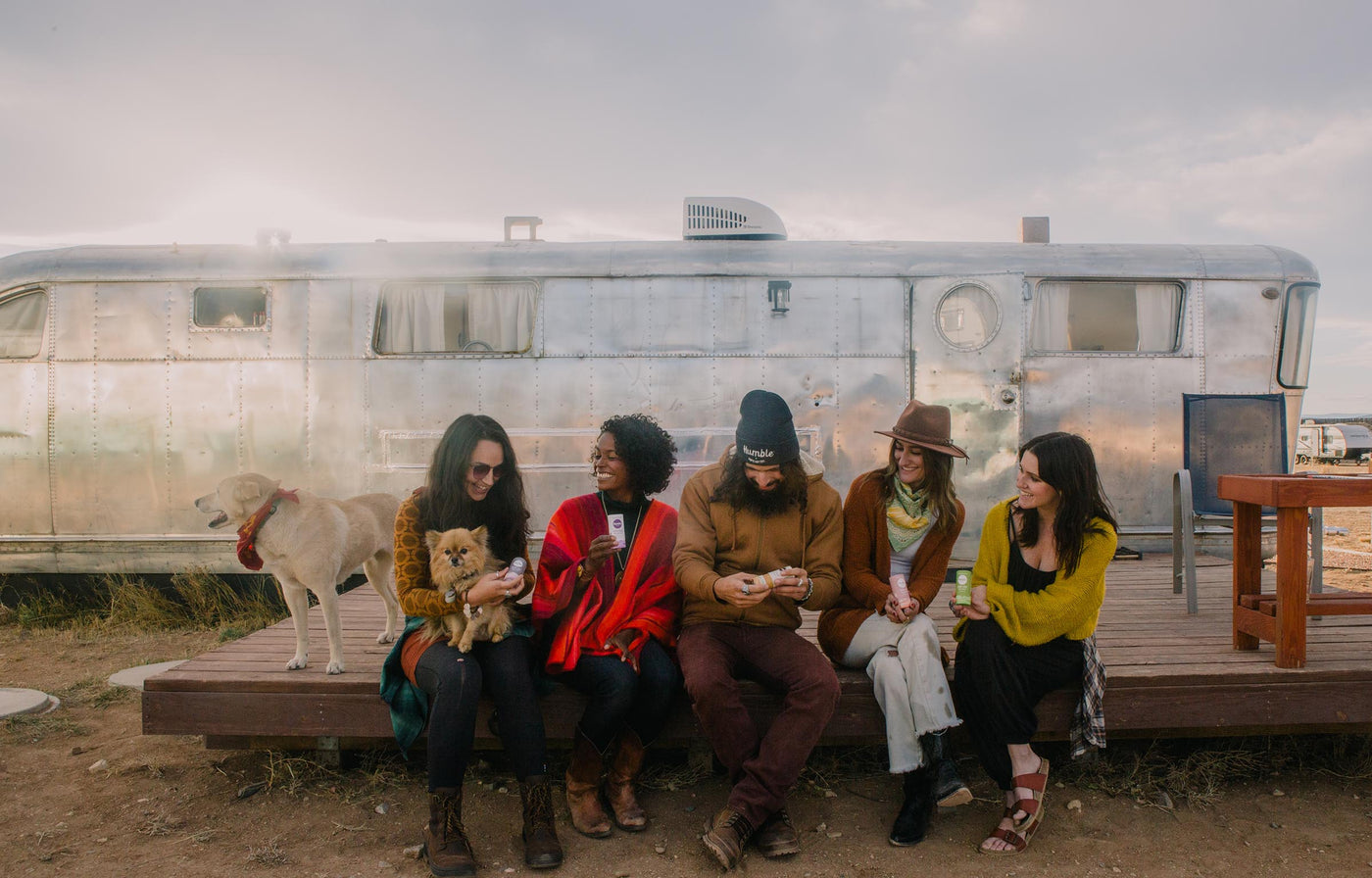 a dog stands with a  group of smiling people sitting on a porch and holding sticks of deodorant 