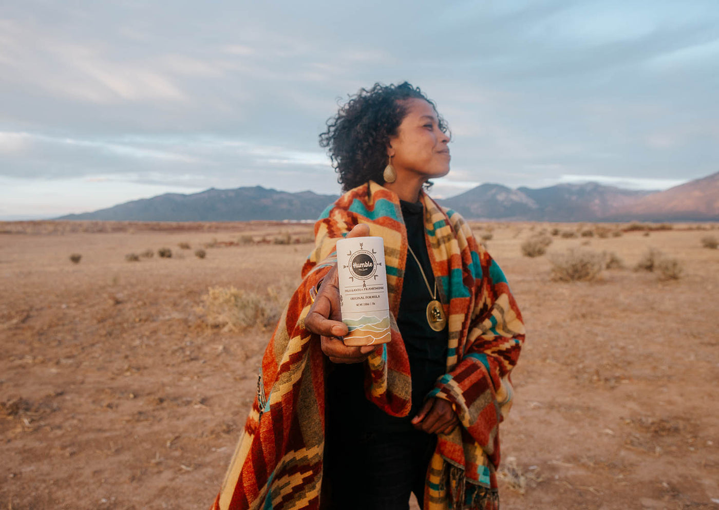 a woman in a colorful coat holds out a deodorant stick with the desert in the background 